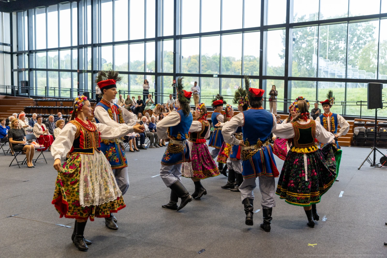 Grupa tancerzy w tradycyjnych strojach ludowych tańczy w sali, widownia obserwuje występ.  Autor: Bogusław Świerzowski