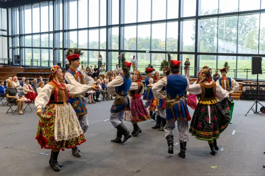 Grupa tancerzy w tradycyjnych strojach ludowych tańczy w sali, widownia obserwuje występ.