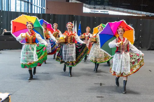 Grupa tancerek w tradycyjnych strojach ludowych z parasolkami w kolorach tęczy.