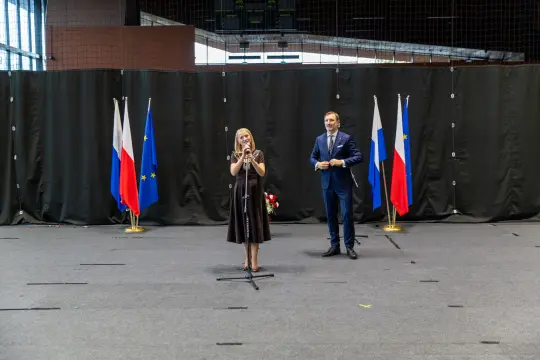 Kobieta w czarnej sukience przemawia na tle flag Polski, Rosji i Unii Europejskiej.
