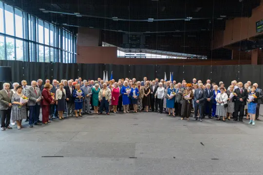 Grupa osób w formalnych strojach, stojących razem w sali, z flagami w tle.