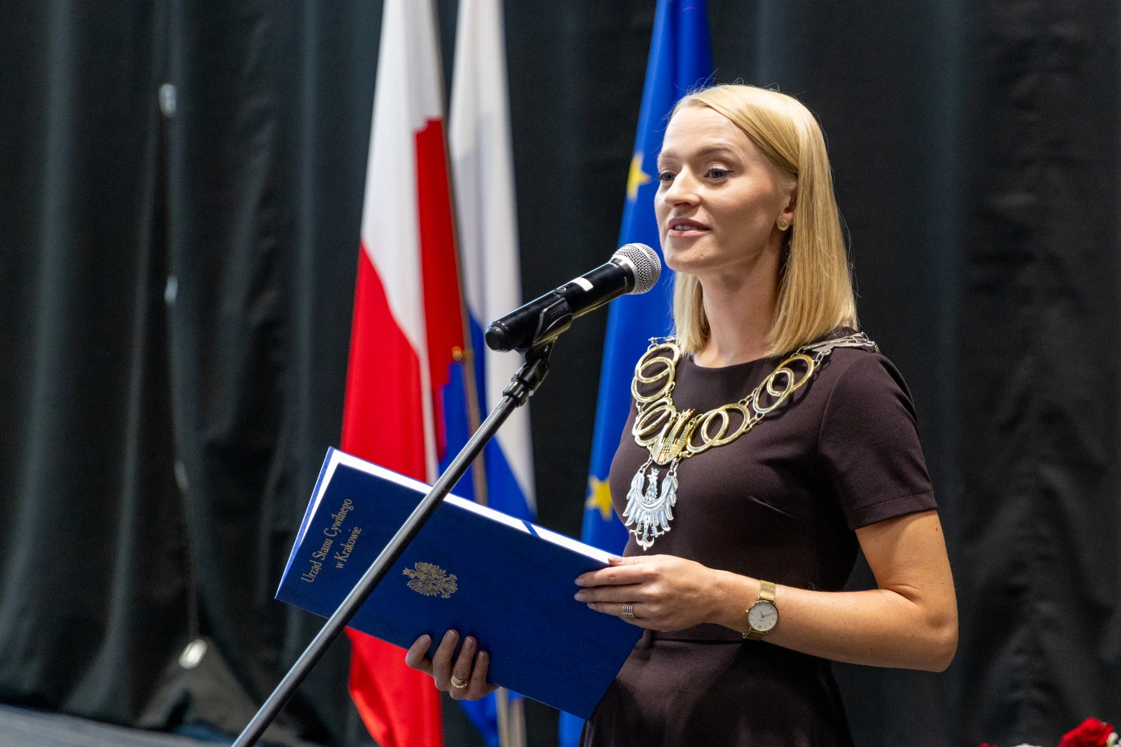 Kobieta przemawia do mikrofonu, trzymając niebieską teczkę, w tle flagi Polski i Unii Europejskiej.  Autor: Bogusław Świerzowski