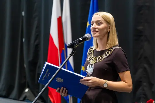 Kobieta przemawia do mikrofonu, trzymając niebieską teczkę, w tle flagi Polski i Unii Europejskiej.