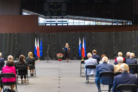 Uroczystość z mówcą na tle flag, zgromadzenie ludzi siedzących na krzesłach.