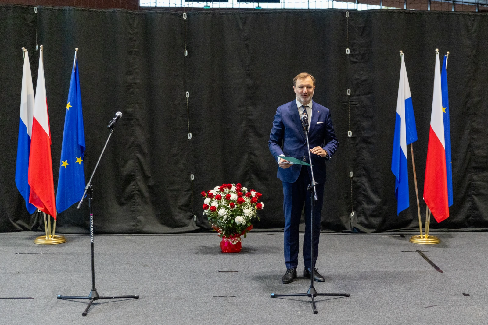 Mężczyzna w garniturze stoi przy mikrofonach, obok flagi Polski i Unii Europejskiej oraz bukietu kwiatów.  Autor: Bogusław Świerzowski