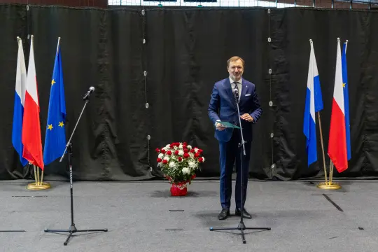 Mężczyzna w garniturze stoi przy mikrofonach, obok flagi Polski i Unii Europejskiej oraz bukietu kwiatów.
