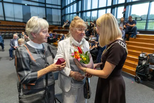 Kobiety z kwiatami i prezentem rozmawiają z uśmiechniętą kobietą w eleganckim stroju.