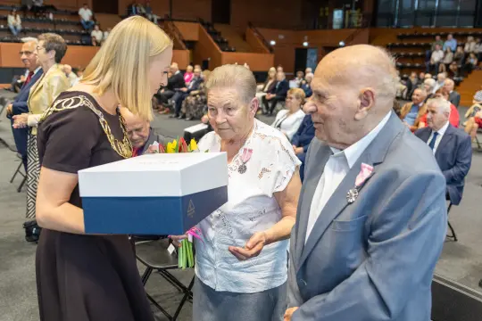 Wręczenie nagrody starszej parze podczas uroczystości z kwiatami i prezentem.