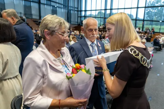 Kobieta z bukietem kwiatów rozmawia z mężczyzną w garniturze podczas uroczystości.