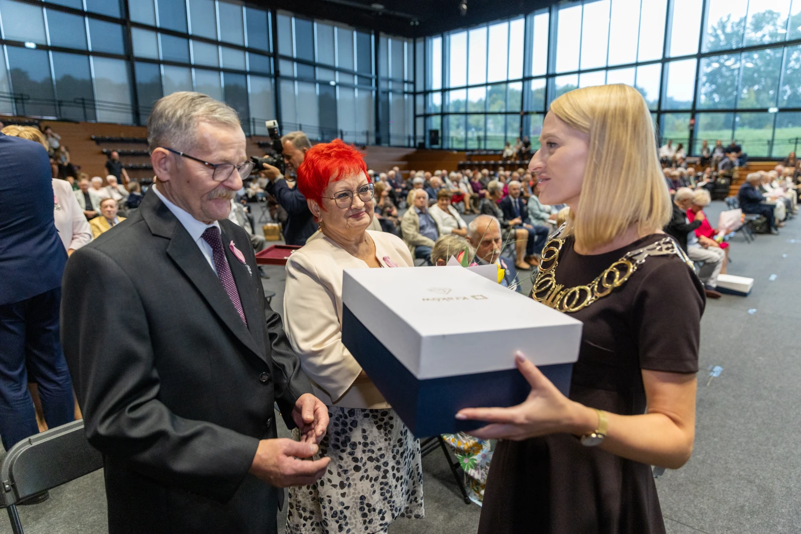 Wręczenie nagrody przez kobietę w eleganckiej sukience mężczyźnie i kobiecie w sali pełnej widzów.  Autor: Bogusław Świerzowski