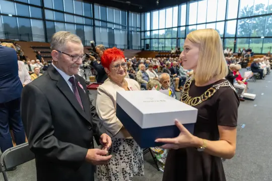 Wręczenie nagrody przez kobietę w eleganckiej sukience mężczyźnie i kobiecie w sali pełnej widzów.