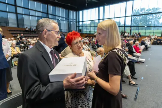 Uroczystość wręczenia nagród dla seniorów w sali z dużymi oknami.