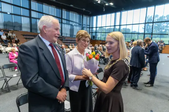 Kobieta wręcza bukiet kwiatów starszym osobom w sali pełnej ludzi.