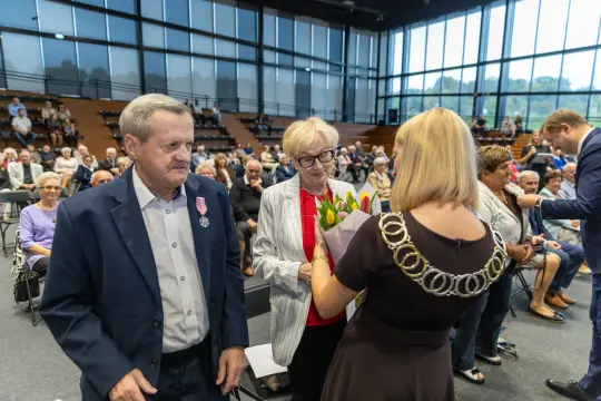 Uroczystość wręczenia nagród z kwiatami i odznaczeniami w tle zgromadzeni goście.