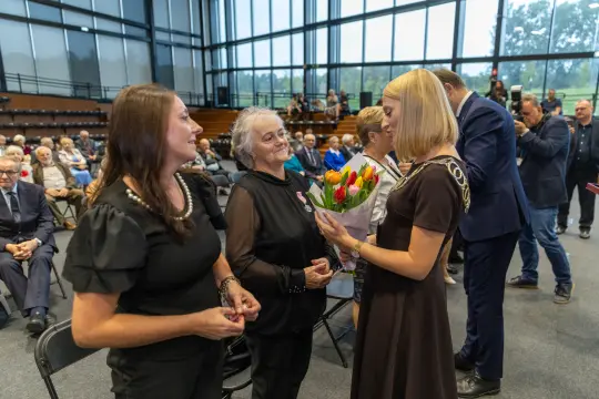 Uroczystość wręczenia kwiatów, obecni goście w tle.