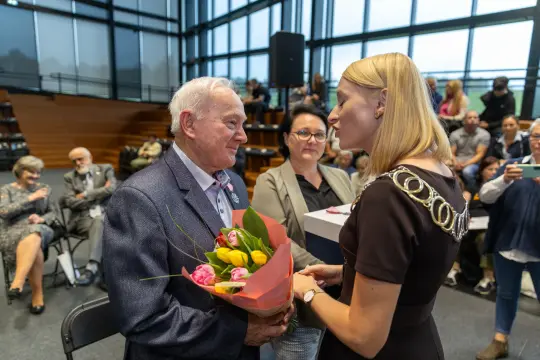 Uroczystość wręczenia kwiatów starszemu mężczyźnie przez kobietę w eleganckim stroju.