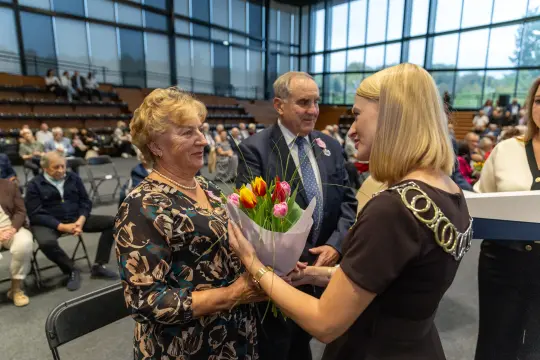 Kobieta otrzymuje bukiet tulipanów od drugiej kobiety w obecności mężczyzny i publiczności.