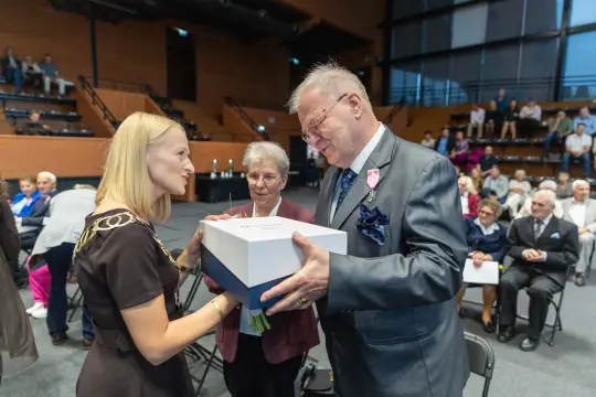 Wręczenie nagrody z okazji uroczystości, obecność gości w tle.