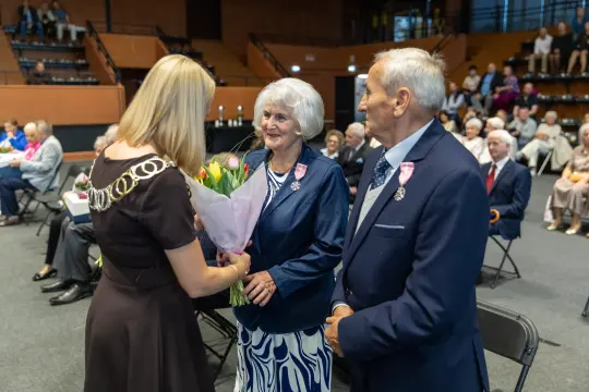 Uroczystość wręczenia kwiatów parze starszych ludzi w sali pełnej gości.