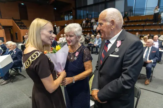 Uroczystość z wręczeniem kwiatów i odznaczeń, uczestnicy w eleganckich strojach.