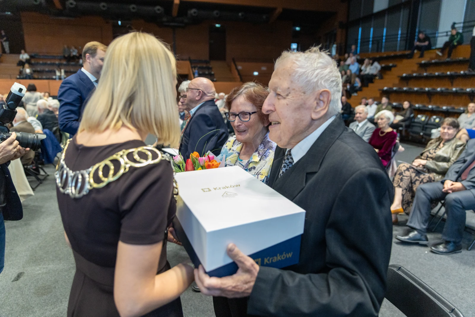 Uroczystość wręczenia nagród z kwiatami i prezentem w tle siedzący goście.  Autor: Bogusław Świerzowski