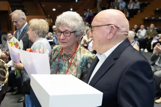 Starsza para z bukietami kwiatów i prezentami, otoczona przez innych uczestników wydarzenia.