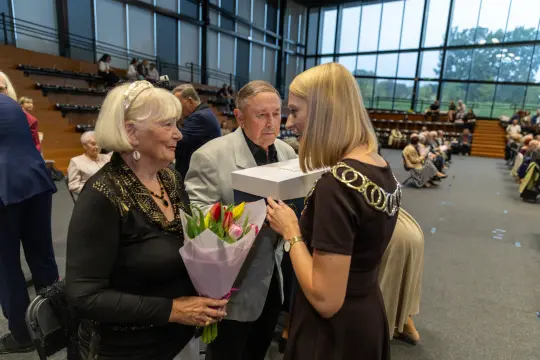 Kobieta z bukietem kwiatów wręcza prezent mężczyźnie w eleganckim otoczeniu.
