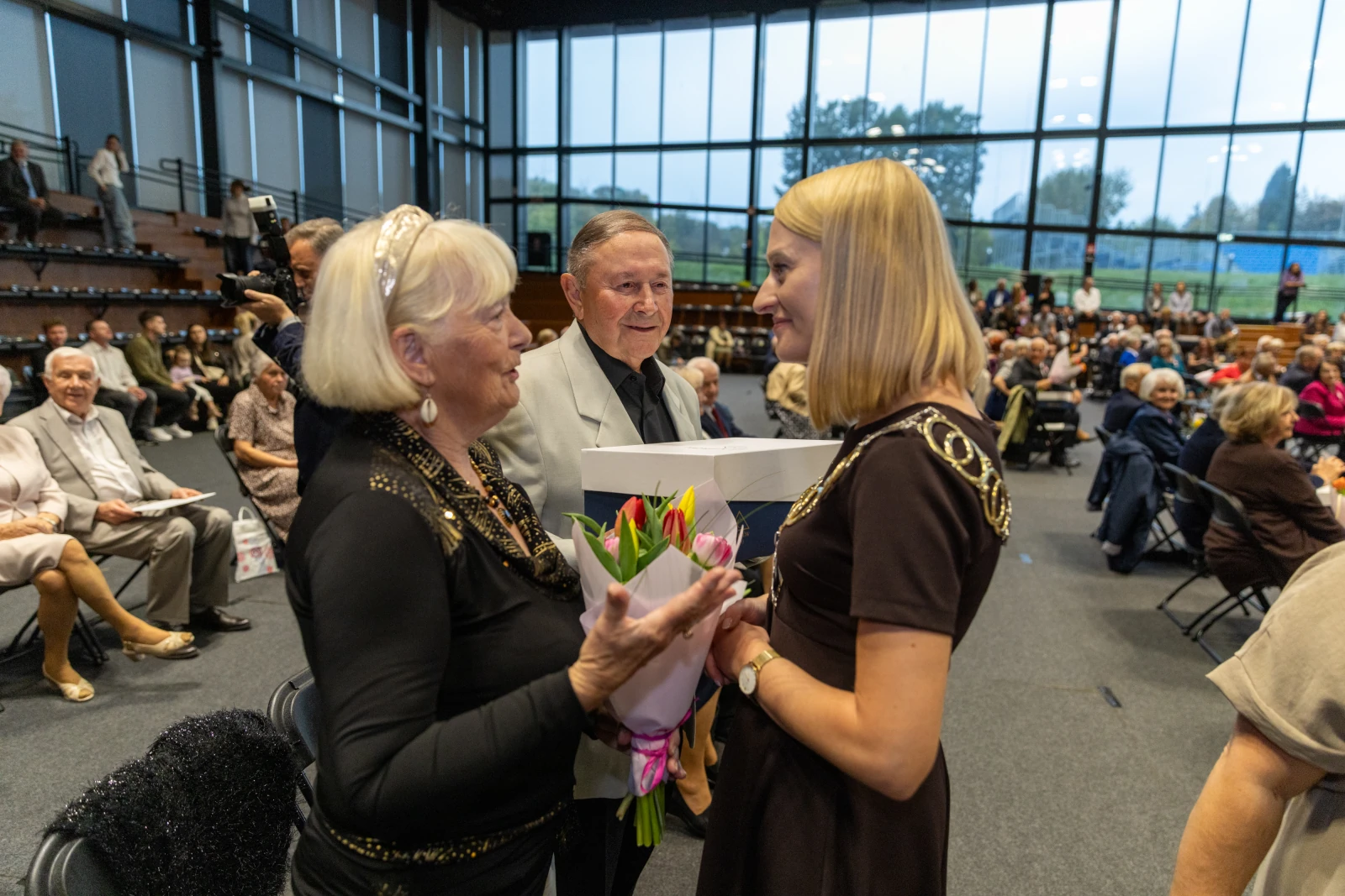 Kobieta wręcza bukiet kwiatów innej kobiecie w towarzystwie starszych osób na sali.  Autor: Bogusław Świerzowski
