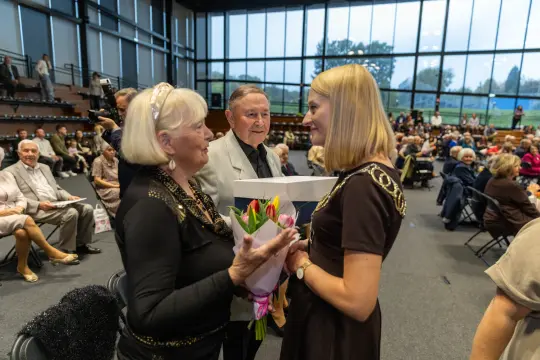 Kobieta wręcza bukiet kwiatów innej kobiecie w towarzystwie starszych osób na sali.