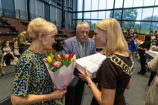 Kobieta z bukietem tulipanów odbiera nagrodę od drugiej kobiety. Mężczyzna obserwuje.