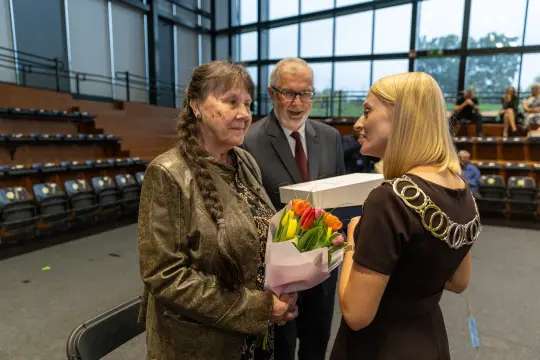 Kobieta z bukietem kwiatów rozmawia z dwojgiem osób w sali.