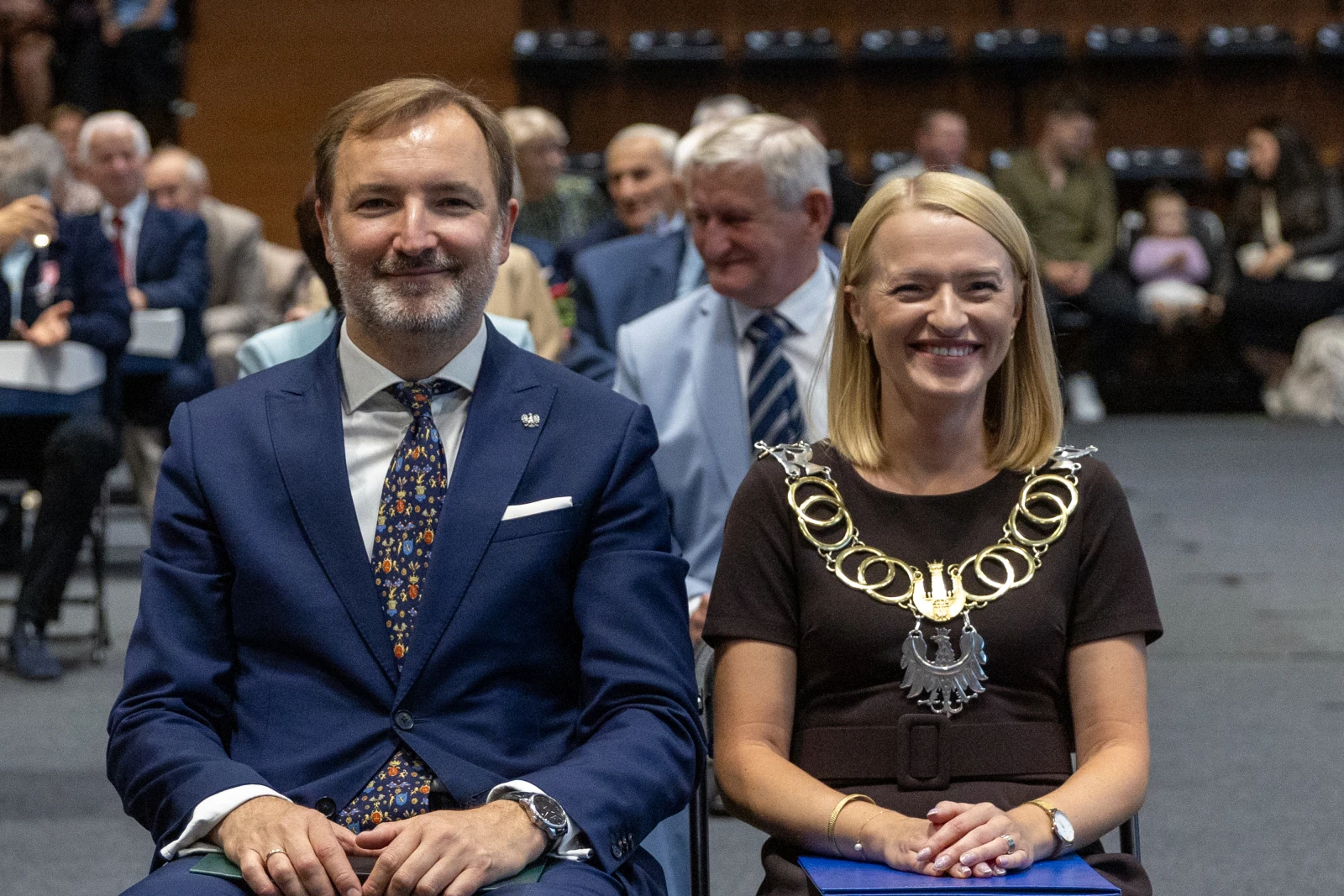 Mężczyzna w garniturze i kobieta w łańcuchu łańcuchu, siedzący na ceremonii wśród publiczności.  Autor: Bogusław Świerzowski