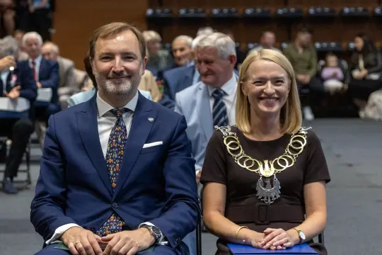 Mężczyzna w garniturze i kobieta w łańcuchu łańcuchu, siedzący na ceremonii wśród publiczności.