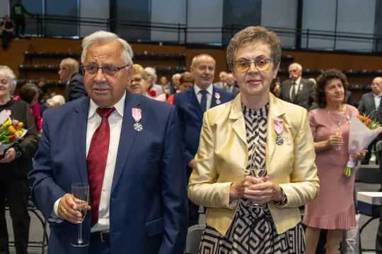 Uroczystość wręczenia odznaczeń, goście w eleganckich strojach, kwiaty w rękach.