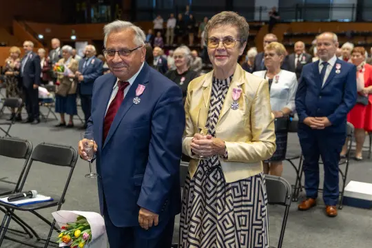 Para w eleganckich strojach, z odznaczeniami, podczas uroczystości w tłumie gości.