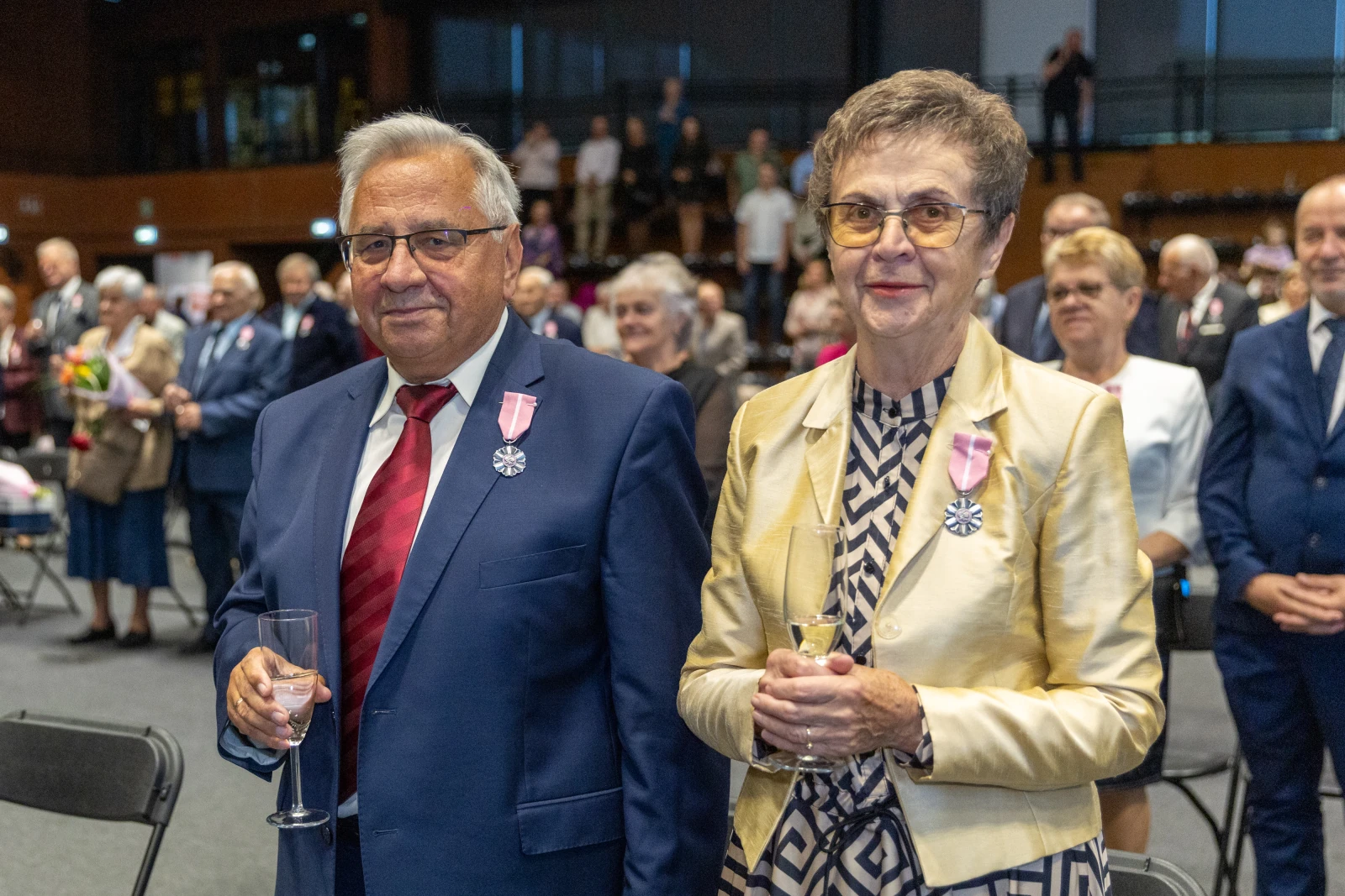 Para osób w eleganckich strojach, trzymająca kieliszki, w tle zgromadzenie ludzi.  Autor: Bogusław Świerzowski