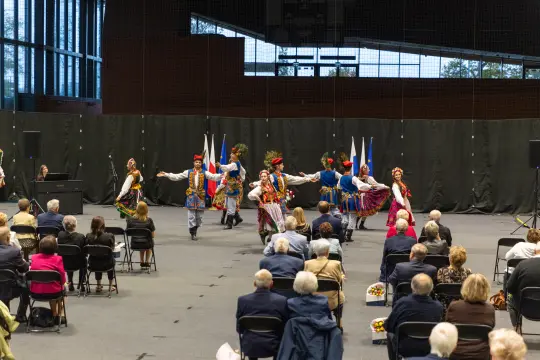 Tancerze w tradycyjnych strojach ludowych wykonują taniec na scenie przed publicznością.