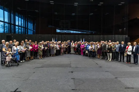 Grupa seniorów stojąca w sali, w tle flagi i ciemne ściany.