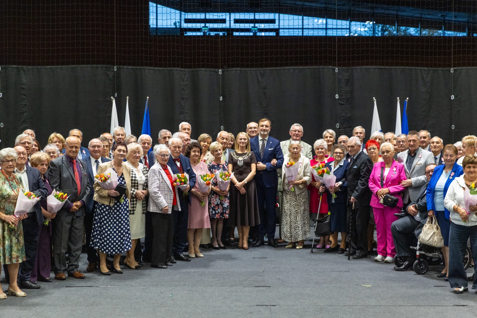 Grupa seniorów z kwiatami, stojąca na tle flag i ciemnej ściany.  Autor: Bogusław Świerzowski