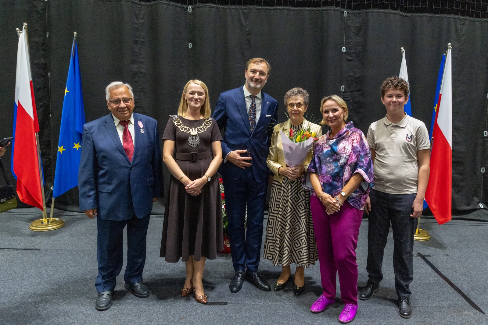 Grupa osób w formalnych strojach, z flagami Polski i Unii Europejskiej w tle.  Autor: Bogusław Świerzowski