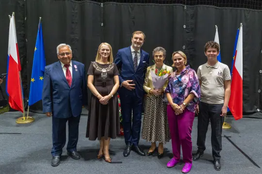 Grupa osób w formalnych strojach, z flagami Polski i Unii Europejskiej w tle.