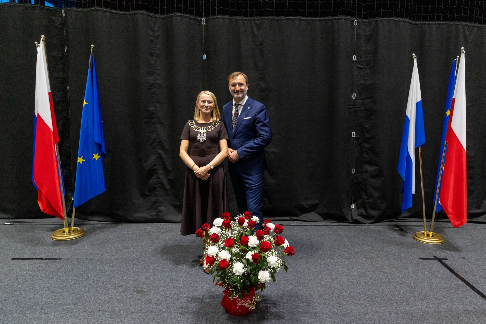 Para w eleganckich strojach stoi obok flag Polski, Unii Europejskiej i Rosji, z bukietem kwiatów.  Autor: Bogusław Świerzowski