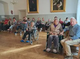 Grupa osób starszych w sali, część na wózkach inwalidzkich, w otoczeniu drewnianych mebli.