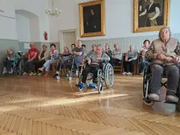 Grupa osób starszych siedzących na wózkach inwalidzkich w sali.