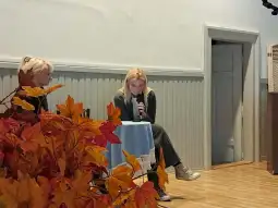 Dwie kobiety siedzą przy stole, jedna z nich czyta tekst, obok kolorowe liście.