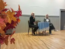 Dwie kobiety siedzą przy stole, jedna z nich mówi do mikrofonu, obok stół z obrusem.