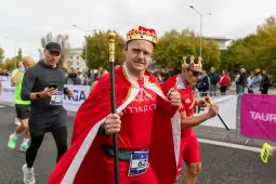 Mężczyzna w koronie i pelerynie biegnie w maratonie, obok innych biegaczy w strojach sportowych.