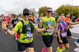 Biegacze w kolorowych strojach uczestniczą w maratonie, uśmiechając się i pokazując znaki zwycięstwa.