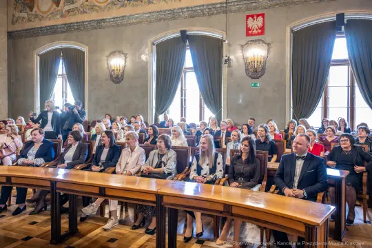 Uczestnicy wydarzenia siedzą w sali z dekoracjami, w tle flaga Polski.