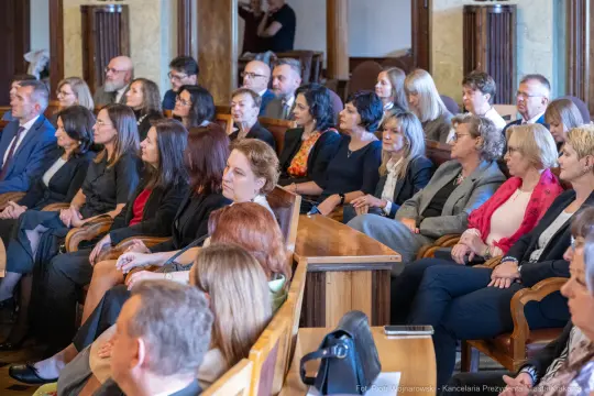 Grupa ludzi siedzących w sali, uczestniczących w wydarzeniu formalnym.